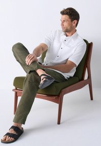 Weißes Kurzarmhemd mit schwarzen Knöpfen, olivgrüne Hose und schwarze Sandalen, sitzt auf einem grünen gepolsterten Holstuhl vor einem weißen Hintergrund.