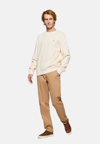 Felpa beige chiaro con colletto rotondo e polsini a costine, abbinata a pantaloni beige e sneakers marroni, esposta su un modello contro uno sfondo neutro.