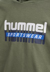 Tissu vert olive avec texte "hummel" en blanc et bleu marine au-dessus d'un rectangle bleu contenant le mot "SPORTSWEAR" en lettres blanches.
