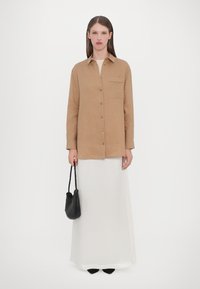 Tan button-up shirt with a chest pocket, paired with a long white skirt and a black woven handbag. Simple design, smooth texture.