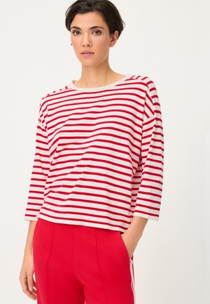 Woman with short dark hair wears a red and white horizontal striped long-sleeve top and red pants, posing with one hand in pocket.