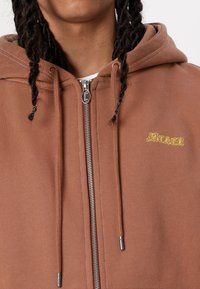 Brown zip-up hoodie made from soft fabric. Features a silver zipper, adjustable drawstrings, and yellow embroidered brand logo on the chest.