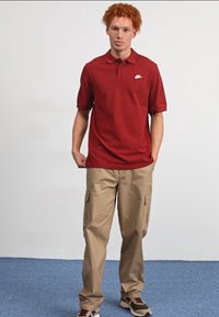 Crvena polo majica s kratkim rukavima i bijelim Nike logom, u kombinaciji sa bež cargo hlačama s bočnim džepovima i smeškim patikama.
