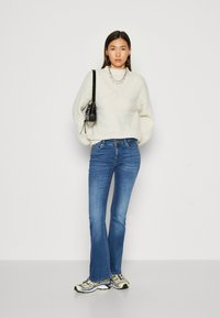 Hvid strikket sweater med rund hals, parret med blå klokkeformede jeans og gule sneakers. Accessories inkluderer en sort taske og lagdelte halskæder.