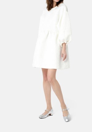 White textured dress with puff sleeves, cinched at the waist. Worn with shiny silver Mary Jane shoes featuring low heels and ankle strap.