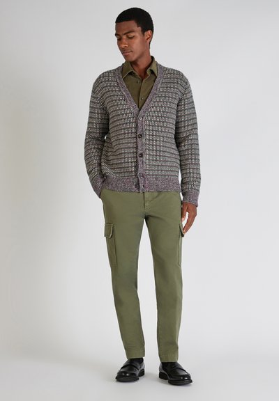 Uomo in piedi che indossa un cardigan grigio con motivo sopra una camicia verde oliva con bottoni, pantaloni cargo verdi oliva e scarpe nere.