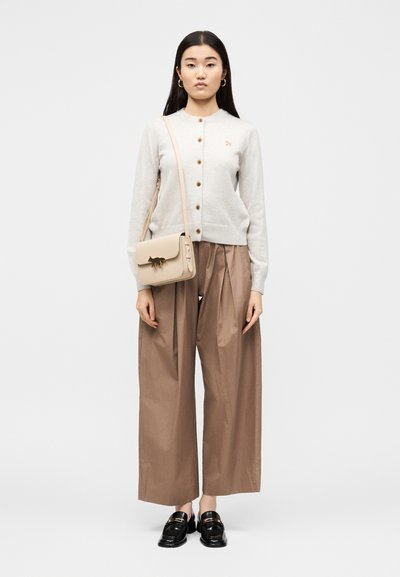 Γυναίκα που φοράει μπεζ cardigan με κουμπιά, φαρδιά καφέ παντελόνια, μαύρα loafers και μπεζ τσάντα χιαστί με έμβλημα γάτας.