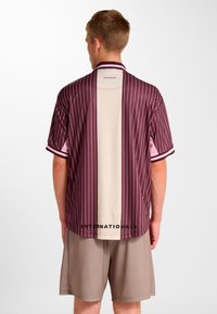 Koszulka z krótkim rękawem w ciemnym bordowym kolorze z pionowymi paskami, z beżowym panelem na środku i czarnym napisem przy krawędzi.