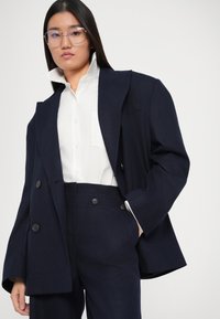 Marineblazer mit doppelt geknöpften Knöpfen, kombiniert mit einem weißen Hemd mit einer Brusttasche und hochtaillierten marineblauen Hosen.