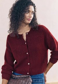Burgunderrød tekstureret cardigan med rund halsudskæring og fem sølvknapper, parret med blå denim. Modellen bærer en grå vævet taske.