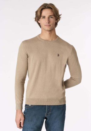 Maglione beige a maglia con collo rotondo, polsini e orlo a costine, con un piccolo logo sul lato sinistro del petto. Indossato con jeans blu in denim.