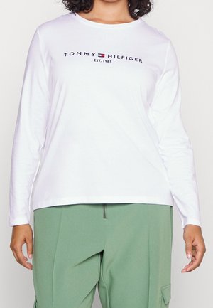 Kobieta w białej koszuli z długim rękawem z logo „Tommy Hilfiger EST. 1985”, w połączeniu z zielonymi spodniami z przednim zamkiem błyskawicznym i patką na kieszeni.