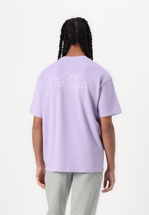 Person med flettet hår set bagfra iført en lyse lilla t-shirt med teksten "Air is Everywhere" og grå bukser.