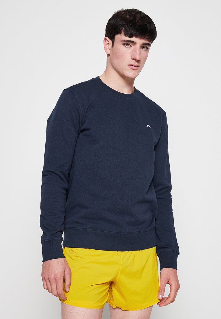 Marineblaue Sweatshirt aus Baumwollmischung, mit rundem Halsausschnitt, langen Ärmeln und kleinem weißen Logo auf der Brust. Getragen mit gelben Shorts.