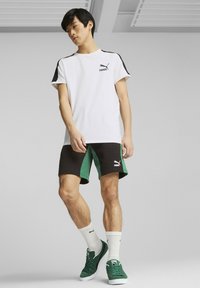 Camiseta blanca de manga corta con detalles en negro, combinada con pantalones cortos negros y verdes. El modelo lleva zapatillas verdes y calcetines blancos con logotipos.