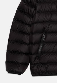 Zwarte pufferjack met gecapitonneerde secties, een zijzak met rits en elastische manchetten, gemaakt van een glad, glanzend materiaal.