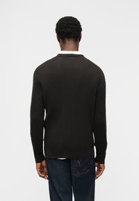 Άνδρας που φοράει μαύρο ριπ πλεκτό πουλόβερ και σκούρο τζιν με κόκκινο κεντητό πουλί στην πίσω τσέπη, στέκεται μπροστά σε απλό λευκό φόντο.