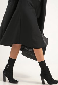 Botines de ante negro con puntera afilada y tacón cuadrado de 7 cm, combinados con una falda negra y calcetas oscuras a juego. Diseño simple y elegante.
