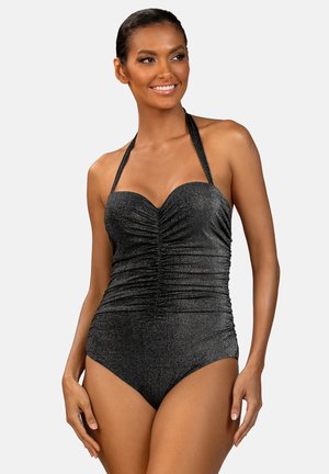 Uśmiechnięta kobieta w marszczonym, błyszczącym czarnym jednoczęściowym stroju kąpielowym na ramiączkach typu halter na jednolitym jasnym tle.