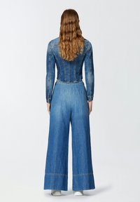 Traper overall s uskim jaknim gornjim dijelom i širokim hlačama. Svijetloplava boja s vidljivim detaljima šivanja i skraćenim dizajnom u struku.