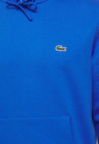 Sweat à capuche bleu avec poche avant et cordons, avec un petit logo crocodile vert brodé sur la poitrine.