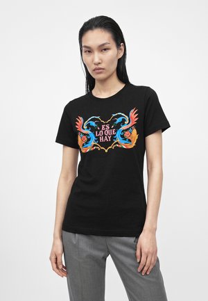 Jeune personne aux cheveux raides et foncés porte un t-shirt noir avec un motif floral coloré et le texte "ES LO QUE HAY", associé à un pantalon gris.