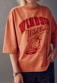 Korallivärvi ülikõrge puuvillane t-särk, millel on punane graafiline kujutis kassist ja kausist. Disain sisaldab julgeid kirjatüüpe ja lõdvestunud istuvust.