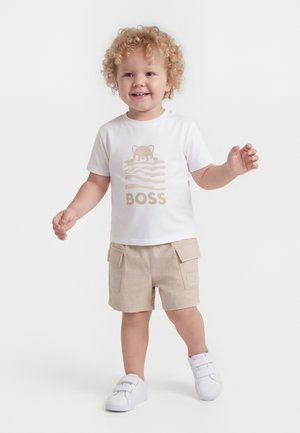 Väikelaps, kellel on lokkis blondid juuksed, valge „BOSS“ t-särgi, beežide taskute ja valgete tossudega, seisab käed kergelt tõstetud.