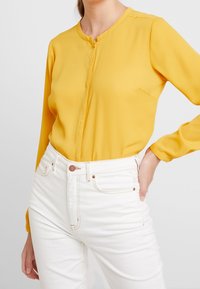 Gelbe Bluse mit rundem Ausschnitt, langen Ärmeln und einer Knopfverschluss vorne. Kombiniert mit hochtaillierten weißen Jeans. Glatte Textur.