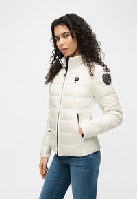 Γυναίκα με σγουρά μαλλιά που φοράει λευκό μπουφάν καπιτονέ με τσέπες με φερμουάρ και μπλε τζιν, στέκεται μπροστά σε απλό ανοιχτό φόντο.