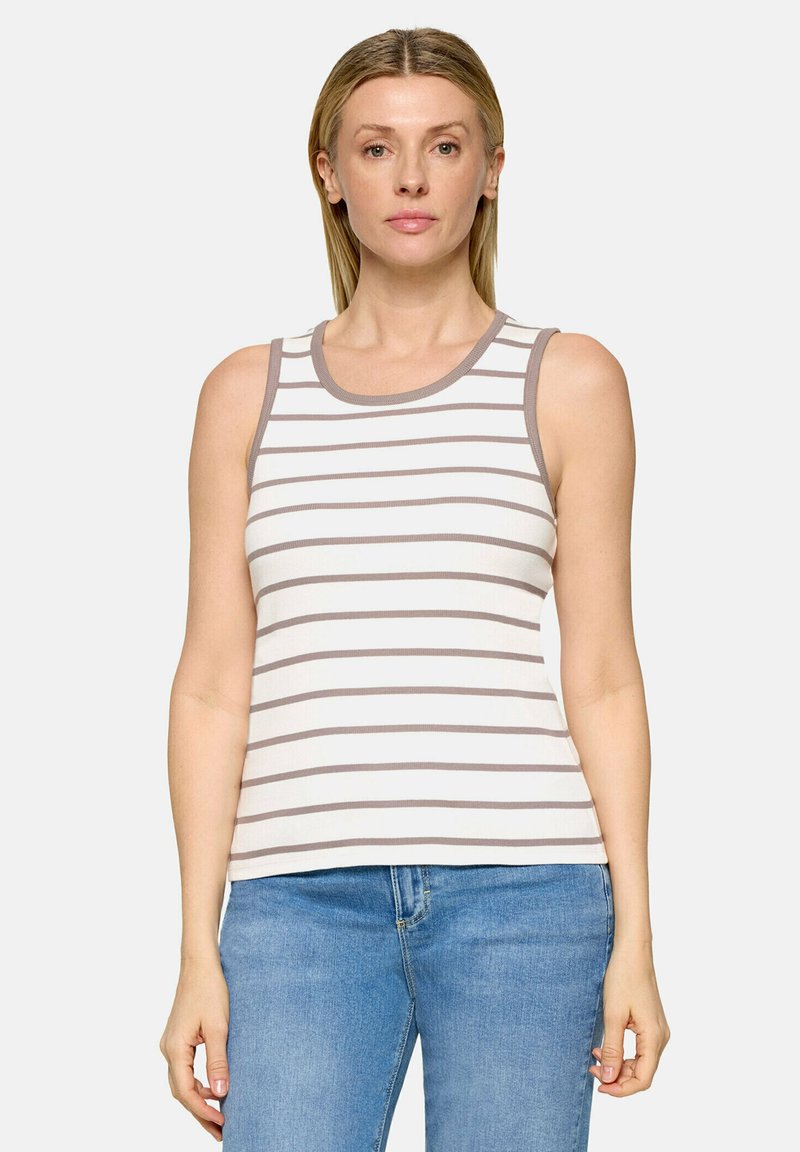 Femme portant un top blanc sans manches avec des rayures horizontales grises et un jean bleu, debout devant un fond uni.
