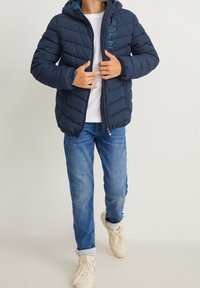 Granatowa, pikowana kurtka z kapturem, z zamkiem błyskawicznym z przodu i detalem z logo. Połączona z niebieskimi dżinsami i jasnymi trampkami.