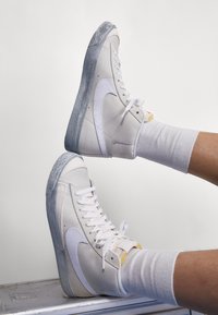 Baskets montantes blanches Nike avec des semelles en gris texturé, portées avec des chaussettes blanches, jambes levées horizontalement contre un fond uni.