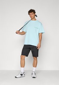 Lyseblå bomull T-skjorte med korte ermer og en liten grønn logo; kombinert med svarte shorts og hvite joggesko med grå detaljer.