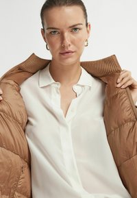 Veste matelassée légère, de couleur marron, avec un col côtelé, portée ouverte sur une chemise blanche à boutons. Texture lisse, design minimaliste.