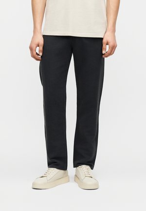 Bas du corps d'une personne portant un pantalon noir à jambes droites, des baskets écrues et une chemise beige sur un fond uni.