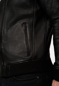 Black leather jacket with a zippered side pocket, ribbed cuffs and hem, and smooth texture. Features a fitted design and subtle stitching.