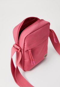 Rosa crossover-veske laget av teksturert stoff, med glidelås, justerbar stropp og frontlomme med dekorative detaljer.