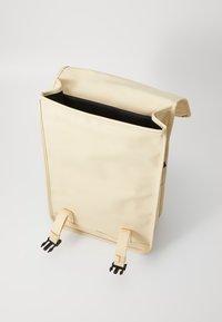 Sac à dos beige à rabat fabriqué en matériau doux, avec un intérieur noir, deux bretelles ajustables et des boucles noires pour une fermeture sécurisée.