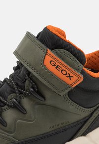 Chaussure imperméable verte avec des accents en tissu noir, arborant un logo orange, une surface texturée, fermée avec des lacets et une sangle à boucle.