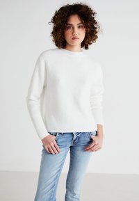 Vit ribbad tröja med rundad halsringning och långa ärmar, kombinerad med ljusblå jeans. Tyget har en mjuk, strukturerad yta.
