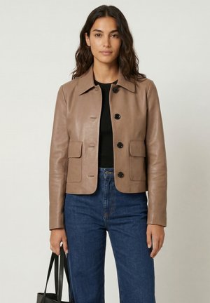 Kvinde med bølget hår iført en tanfarvet læderjakke over en sort top, blå jeans og holder en sort håndtaske mod en ensfarvet baggrund.