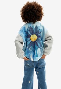 Maglione lavorato a maglia grigio e blu con un grande design di margherita sulla schiena. Abbinato a jeans blu con piccole ricami di fiori bianchi.