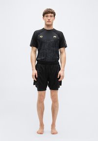 Μαύρο αθλητικό σύνολο που αποτελείται από μια εφαρμοστή κοντομάνικη μπλούζα με υφασμένο σχέδιο και σορτς, και τα δύο με διακριτικές χρυσαφένιες λεπτομέρειες.