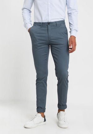 Mężczyzna ubrany w niebieskie spodnie slim fit, białe trampki oraz jasnobłękitną, w paski, koszulę z długim rękawem, z rękami w kieszeniach.