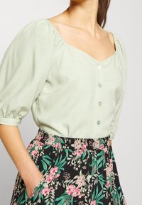 Blouse vert clair avec des manches bouffantes et un décolleté carré, ornée de boutons à l'avant. Associée à une jupe noire à imprimé floral.