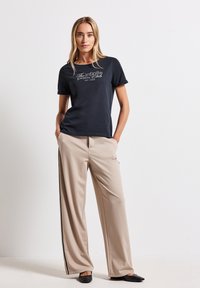 T-shirt marine avec le texte blanc "The infinite Palm Club" et un pantalon large beige avec des rayures noires sur les côtés. Chaussures plates noires.