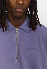 Paarse zip-up shirt met een geribde kraag, zilveren rits en "REVOLUTION" geborduurd op de borst. Zachte katoentextuur.