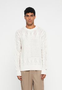Tömör fehér kötött pulóver, különböző fonásmintákkal, textúrált részekkel és bordás aljjal. Hosszú ujjakkal és kerek nyakkivágással tervezték.