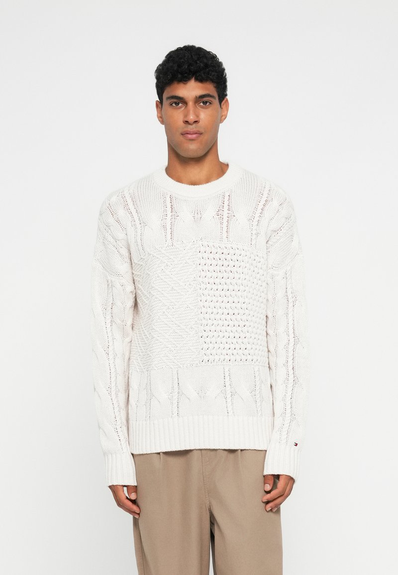 Tömör fehér kötött pulóver, különböző fonásmintákkal, textúrált részekkel és bordás aljjal. Hosszú ujjakkal és kerek nyakkivágással tervezték.